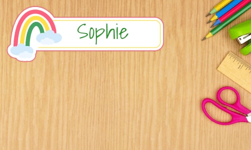 Grade-school desk with colorful name tag sticker that says Sophie with a rainbow graphic in the upper left. Colored pencils, ruler and a pair of pink scissors, green stapler also sit on the right hand side of the desk.