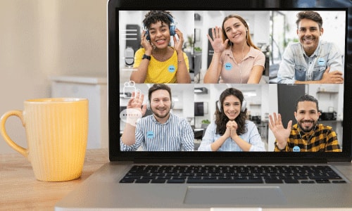 Mac laptop on a desk next to a cnary yellow coffee mug. The computer screen shows six people on a zoom call all wearing name tags.
