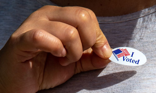 Hand holding an I Voted sticker about to be place on a grey t-shit.