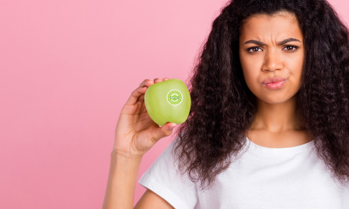 Sark haired young woman in white t-shirt holding a green apple with a fruit sticker on it. Pink background.