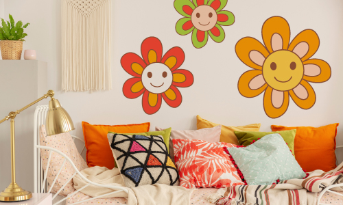 1970's style room with orange, green and teal pillows on a white couch. To the left there is a gold bankers lamp on a table.. There are threee wall stickers on the wall above the couch. They are orange, red-orange and green flowers with happy faces in the center.