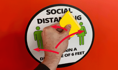 Red wall with social distancing sticker on it. Human male hand is using yellow sticker application squeegee in a zig-zag motion to flatten and help adhere the sticker.