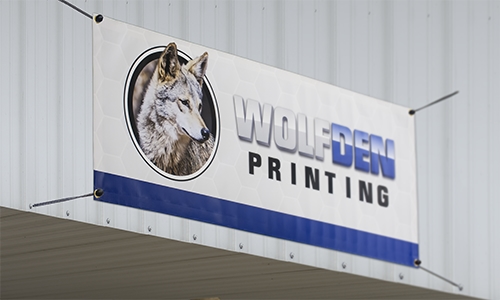 Single-Sided 13-oz. vinyl banner in white with blue border at the bottom. The left side features a company circle logo of a gray wolf head. The text says Wolf Den Printing.